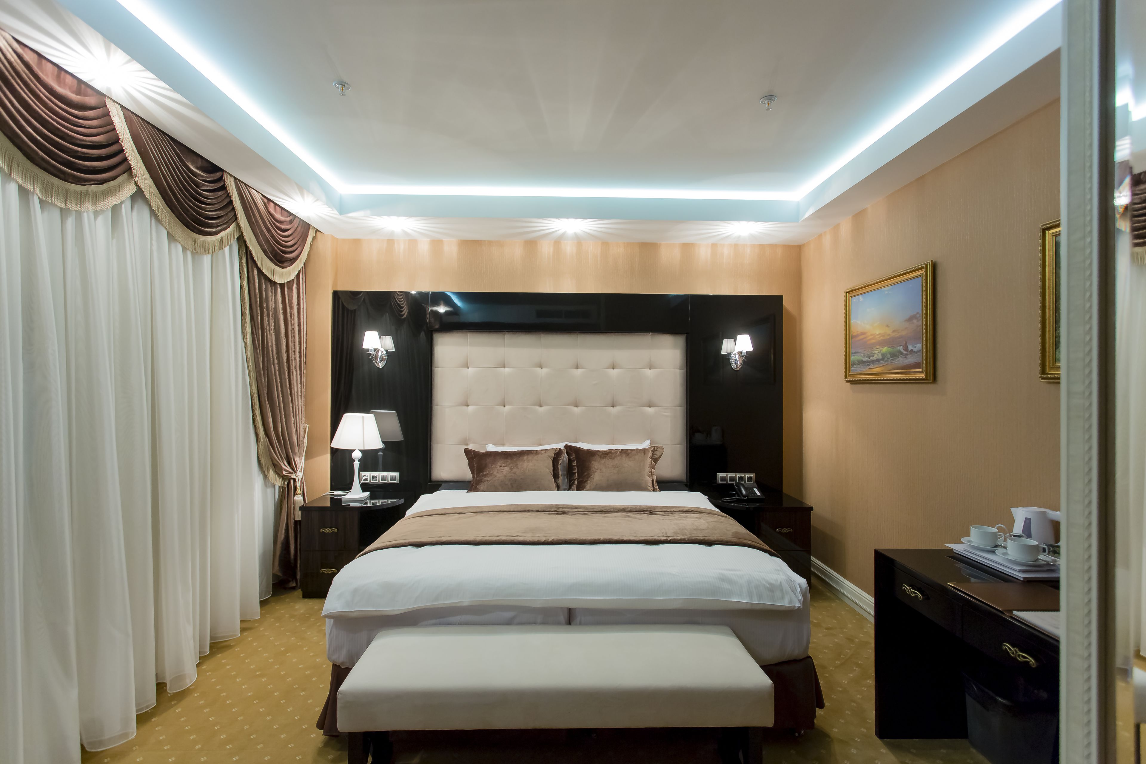 Hotelkamer met een groot bed, hoofdeinde, nachtkastjes, een bankje, gordijnen, een schilderij en een bureau met lamp.