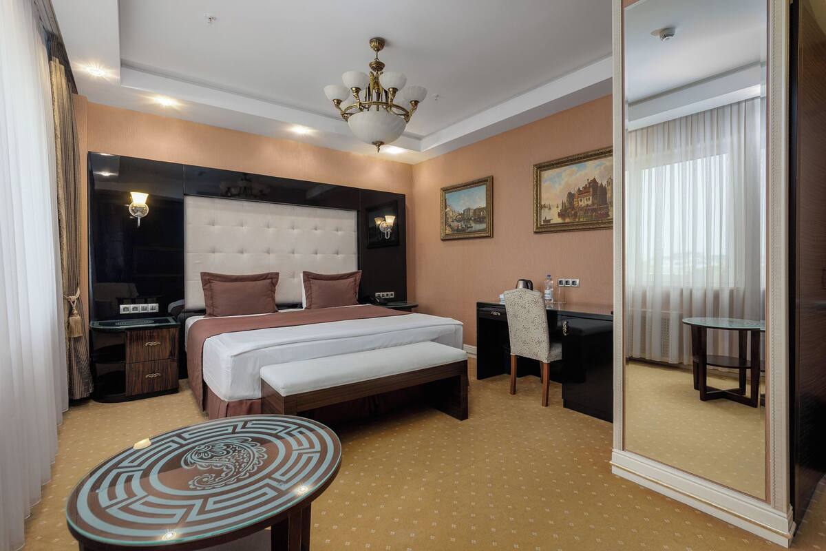 A hotel room with a large bed, a desk, a chair, a small table, a mirror, and a chandelier.