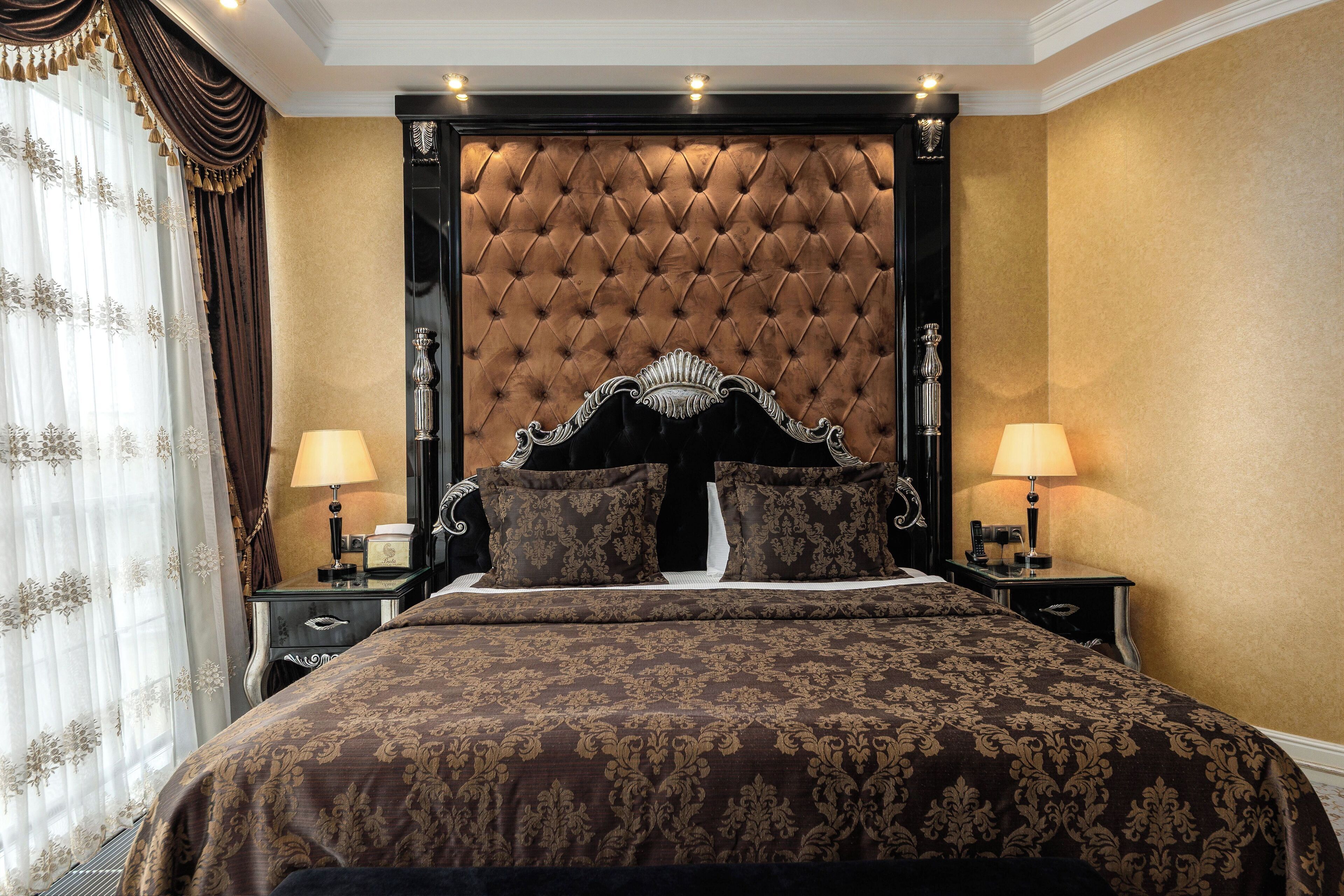 Una cama grande con cabecera capitonada, dos mesitas de noche con lámparas y una ventana con cortinas.