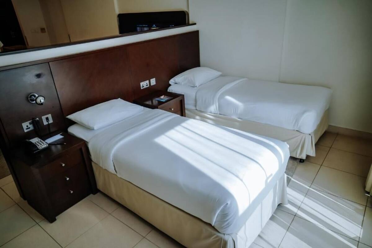 A hotel room with two single beds, a wooden headboard, a bedside table with a phone and a book, and a small nightstand.