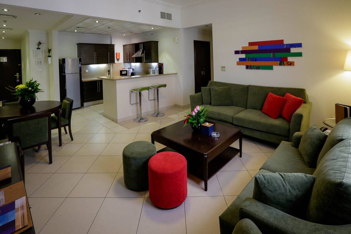 A modern living room with a green sofa, a red ottoman, and a coffee table. There is a kitchen area in the background with a refrigerator, microwave, and bar stools.