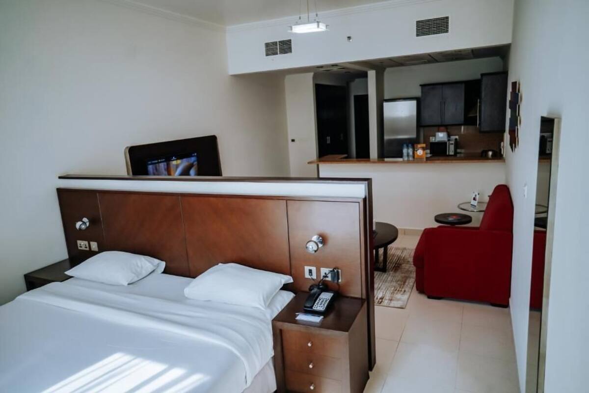 A hotel room with a bed, bedside table, a red armchair, and a kitchenette in the background.