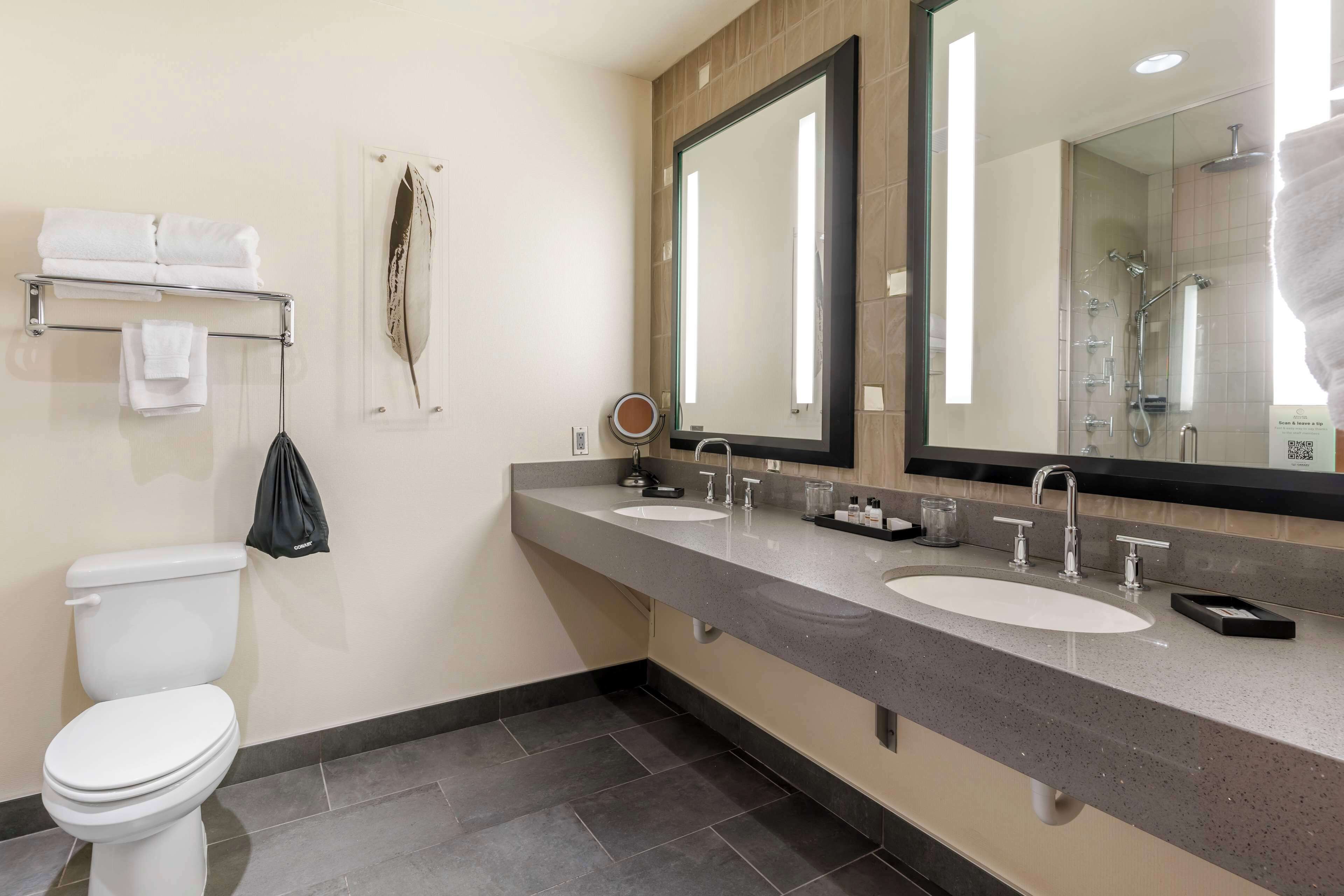 A modern bathroom with a toilet, two sinks, a large mirror, and a towel rack.