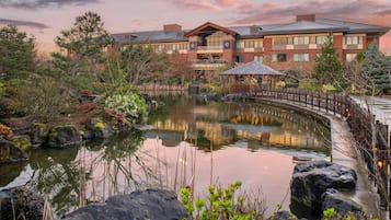 Escape to a serene oasis with a tranquil pond reflecting the hotel's architecture, a wooden bridge leading to a peaceful gazebo.