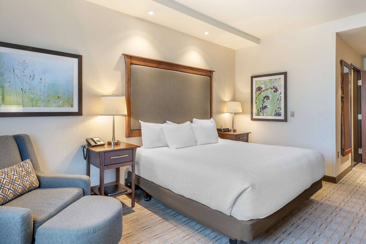 A hotel room with a large bed, a chair, a desk with a lamp, and two wall-mounted lamps.