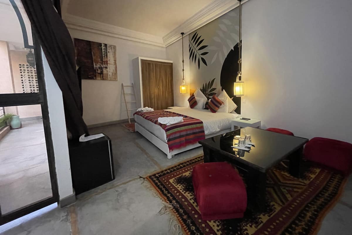 Quarto de hotel moderno com cama, uma mesinha, um banquinho vermelho e uma janela grande com cortinas.