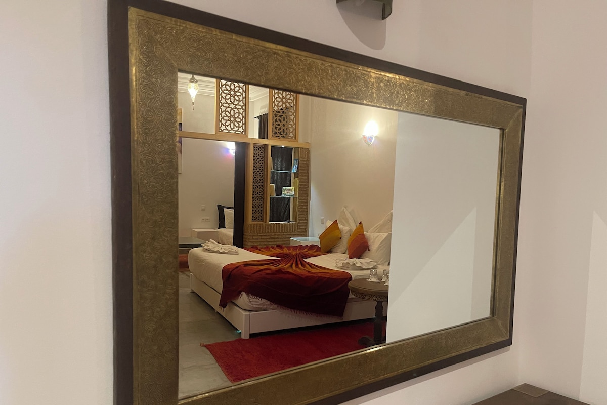 A bedroom with a large mirror, a bed with white bedding and a red throw, a nightstand, and a small table with glasses.