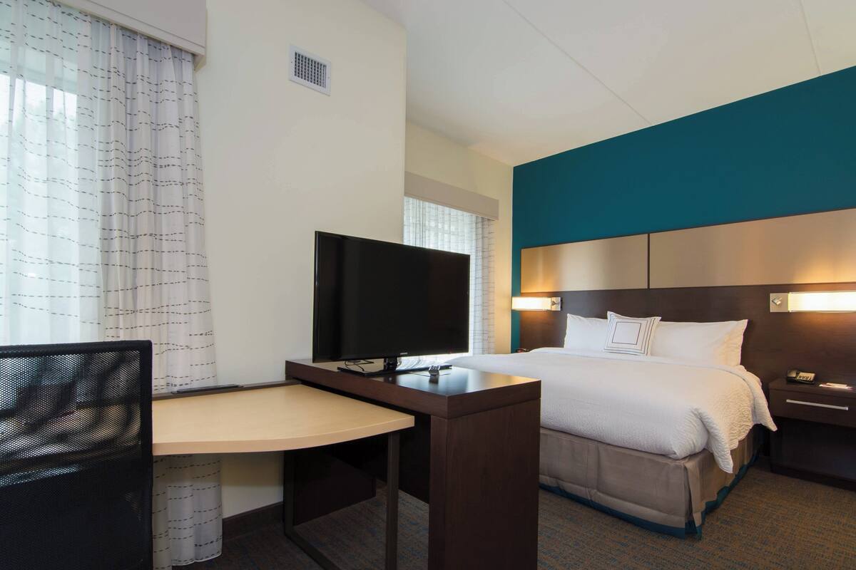A hotel room with a bed, a desk with a TV, a chair, and a bedside table.