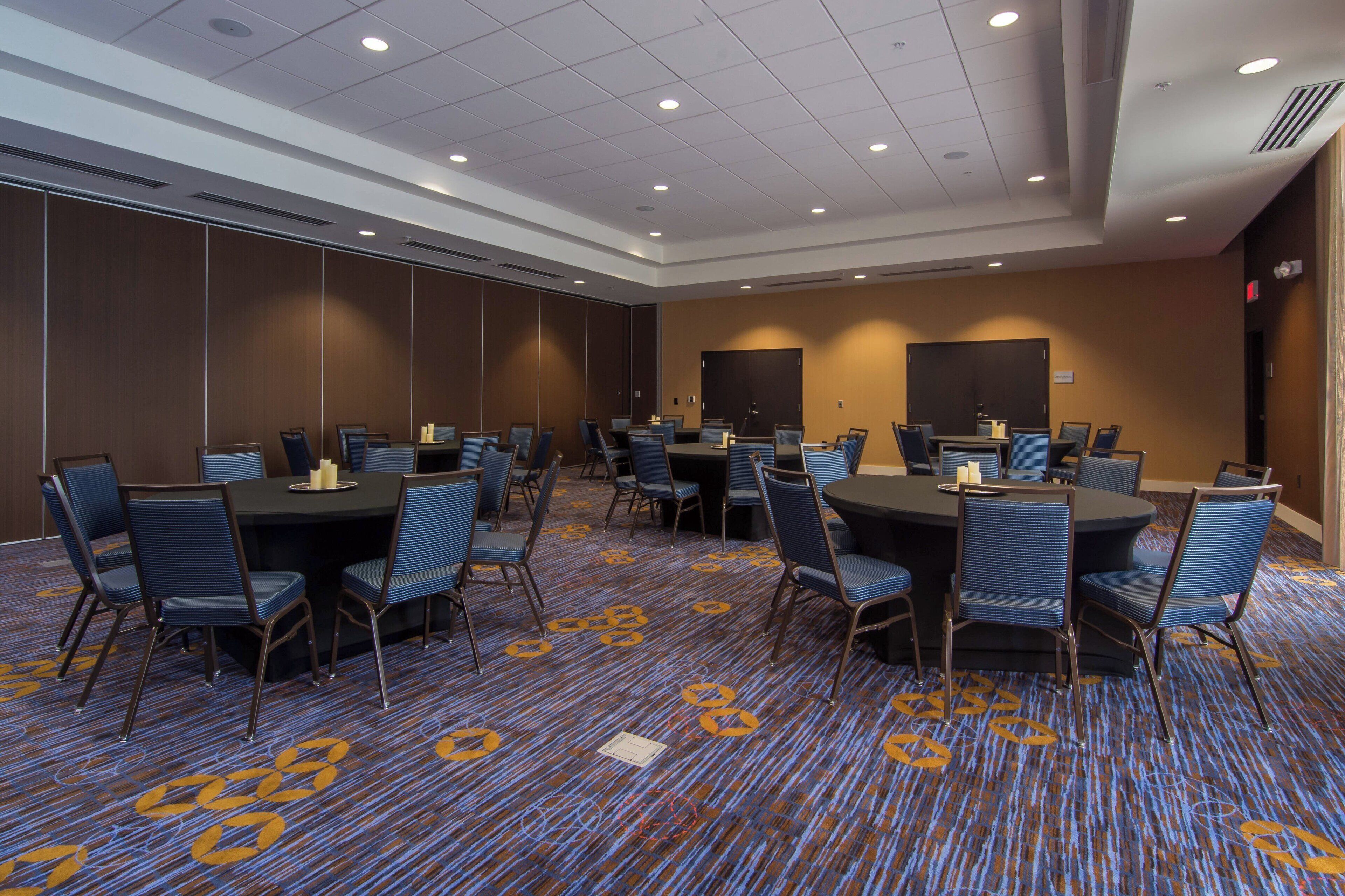 A conference room with multiple tables and chairs, a patterned carpet, and a large screen on the wall.