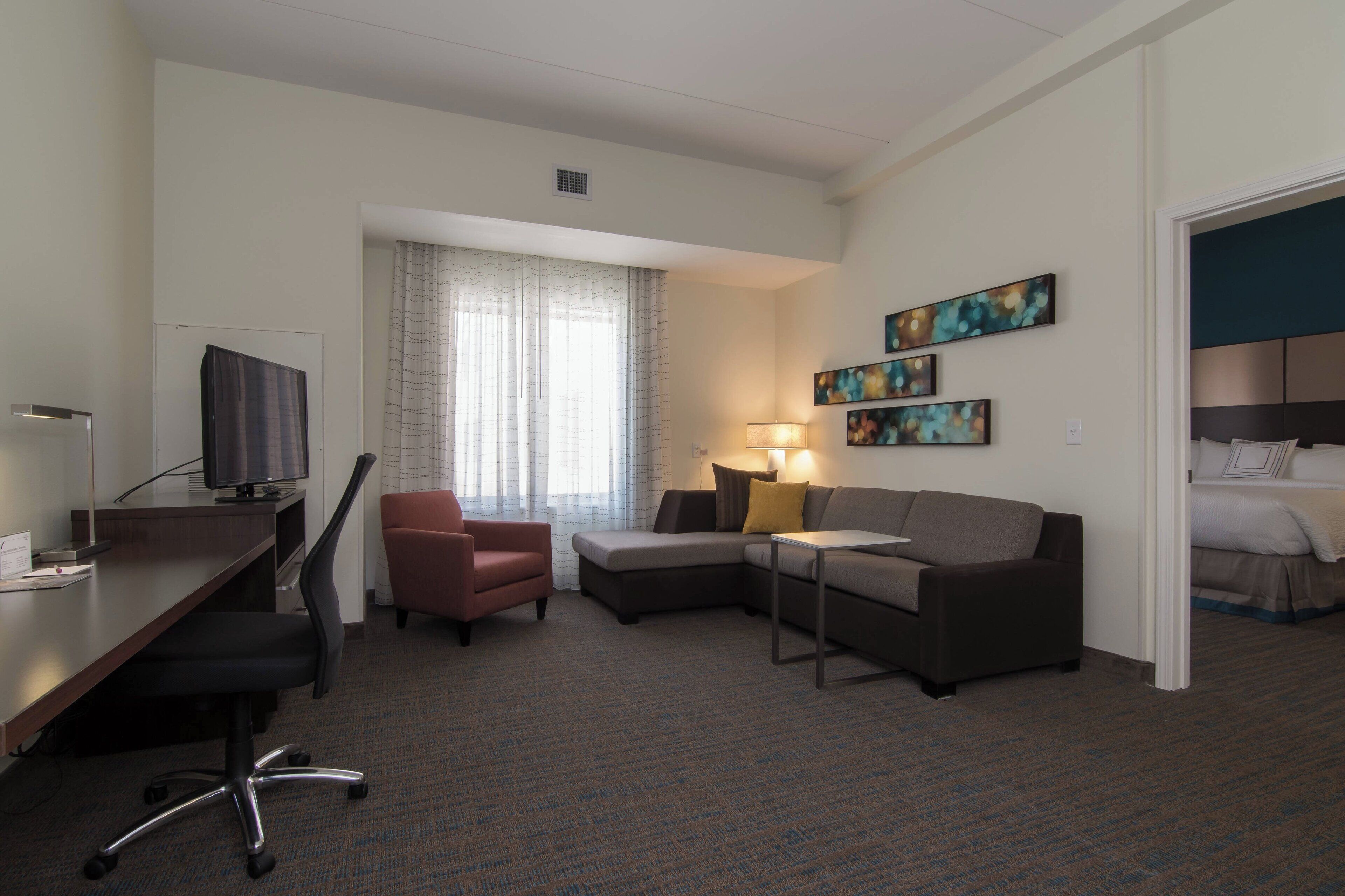 Ein Hotelzimmer mit Schreibtisch, Bürostuhl, Sofa und einem Bett. An der Wand hängen drei Bilder und ein Fenster mit Vorhängen.