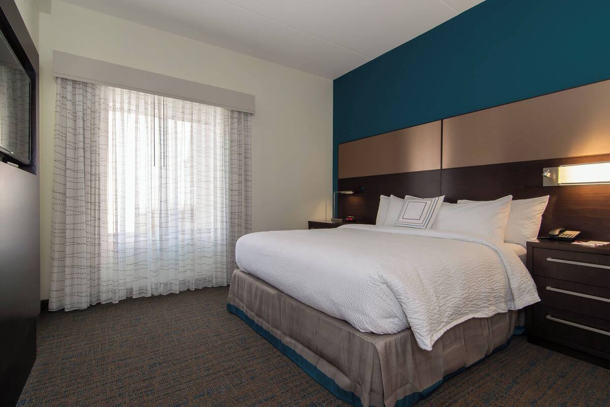 A hotel room with a large bed, a nightstand with a lamp, and a window with sheer curtains.