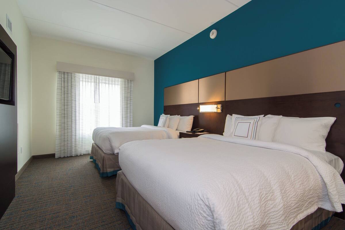 A hotel room with two beds, a large window with curtains, a wall-mounted TV, and a headboard with a light fixture.