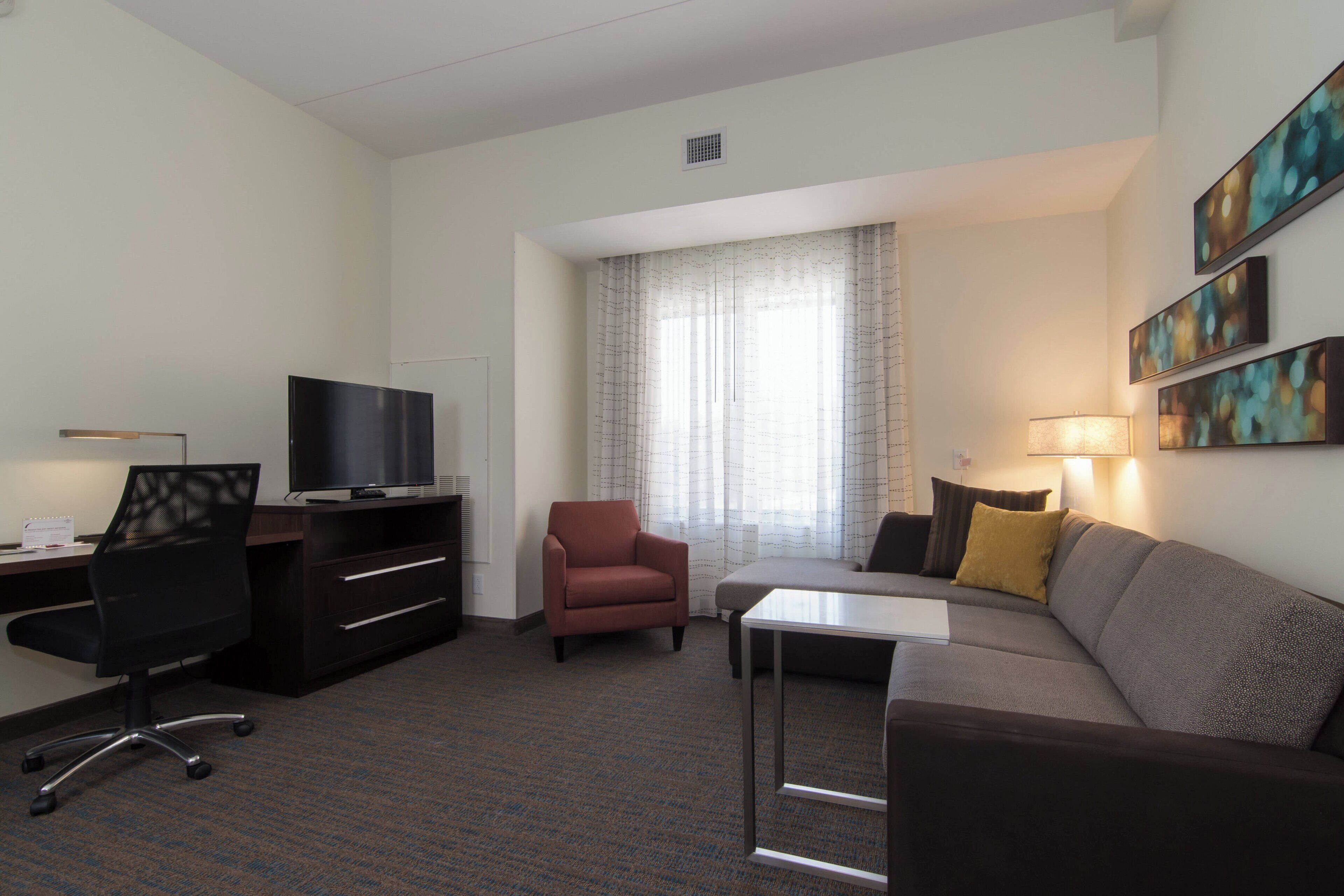 A hotel room with a desk, chair, sofa, TV, and artwork on the wall.