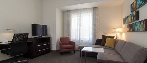 A hotel room with a desk, chair, sofa, TV, and artwork on the wall.