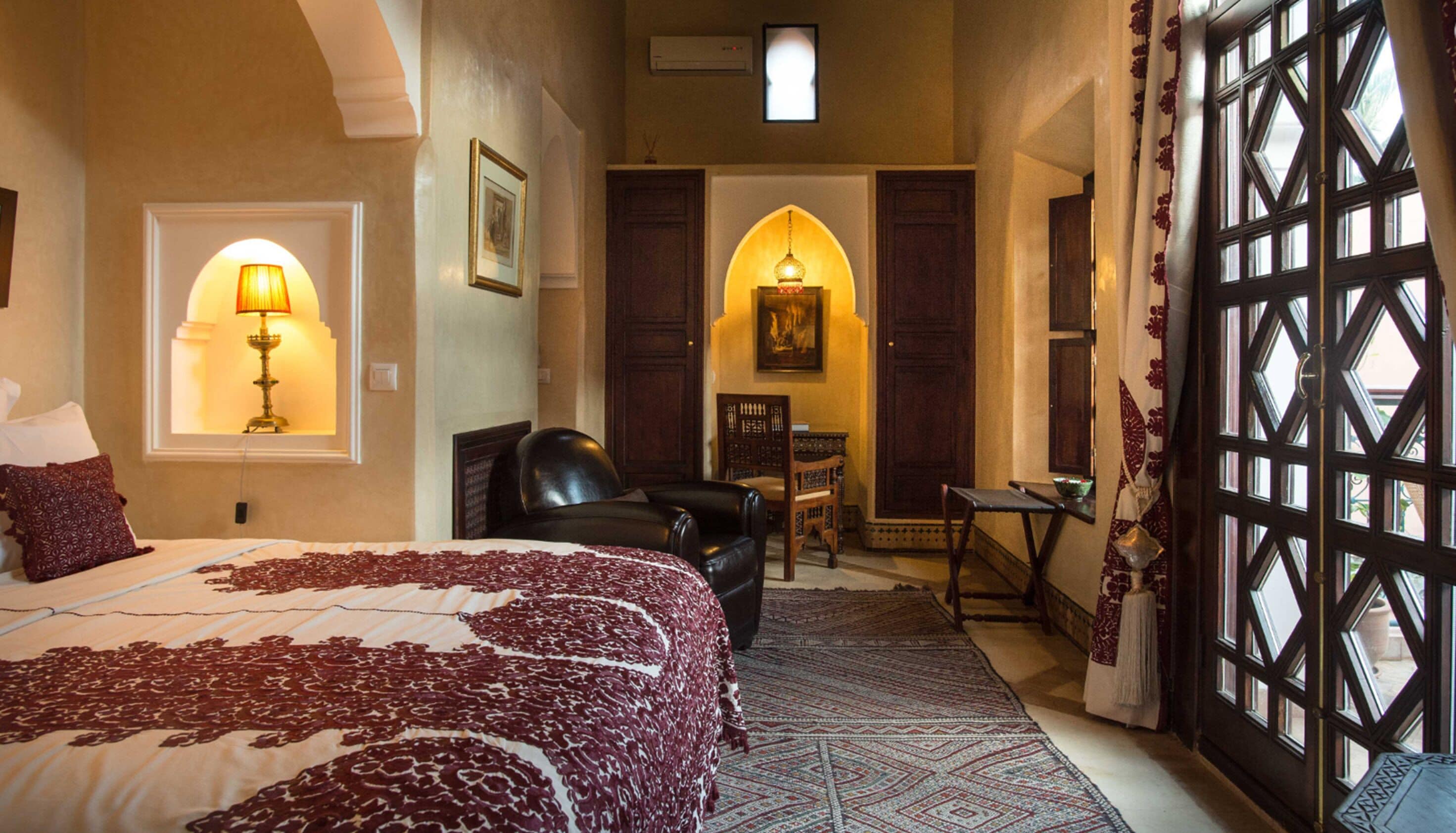 A hotel room with a large bed, a desk, a chair, and a view through a window with a patterned curtain.