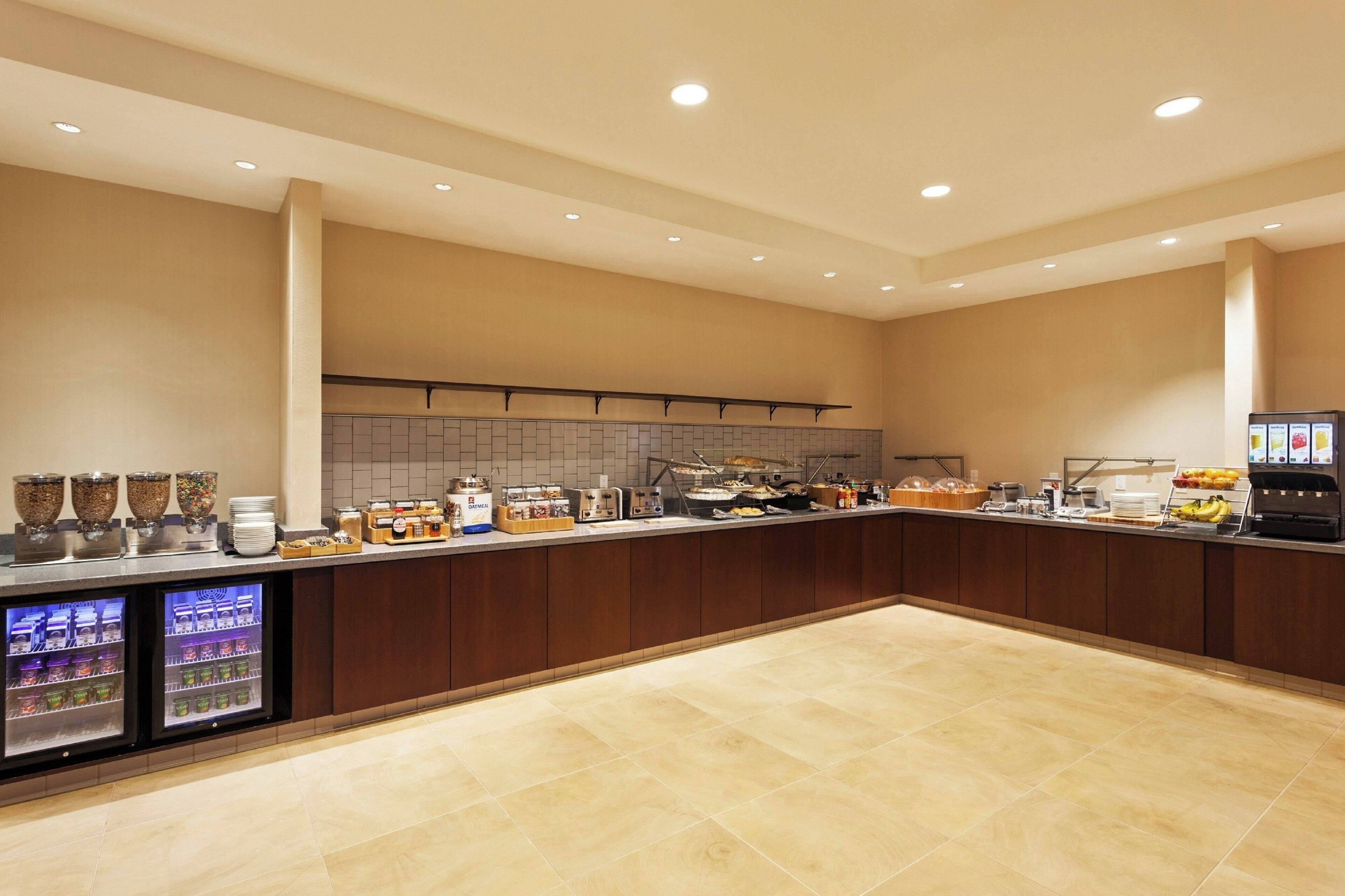 A spacious, well-lit breakfast area with a long counter, refrigerated beverage dispensers, and a variety of food and drink options.