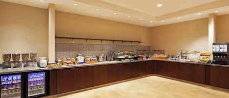 A spacious, well-lit breakfast area with a long counter, refrigerated beverage dispensers, and a variety of food and drink options.
