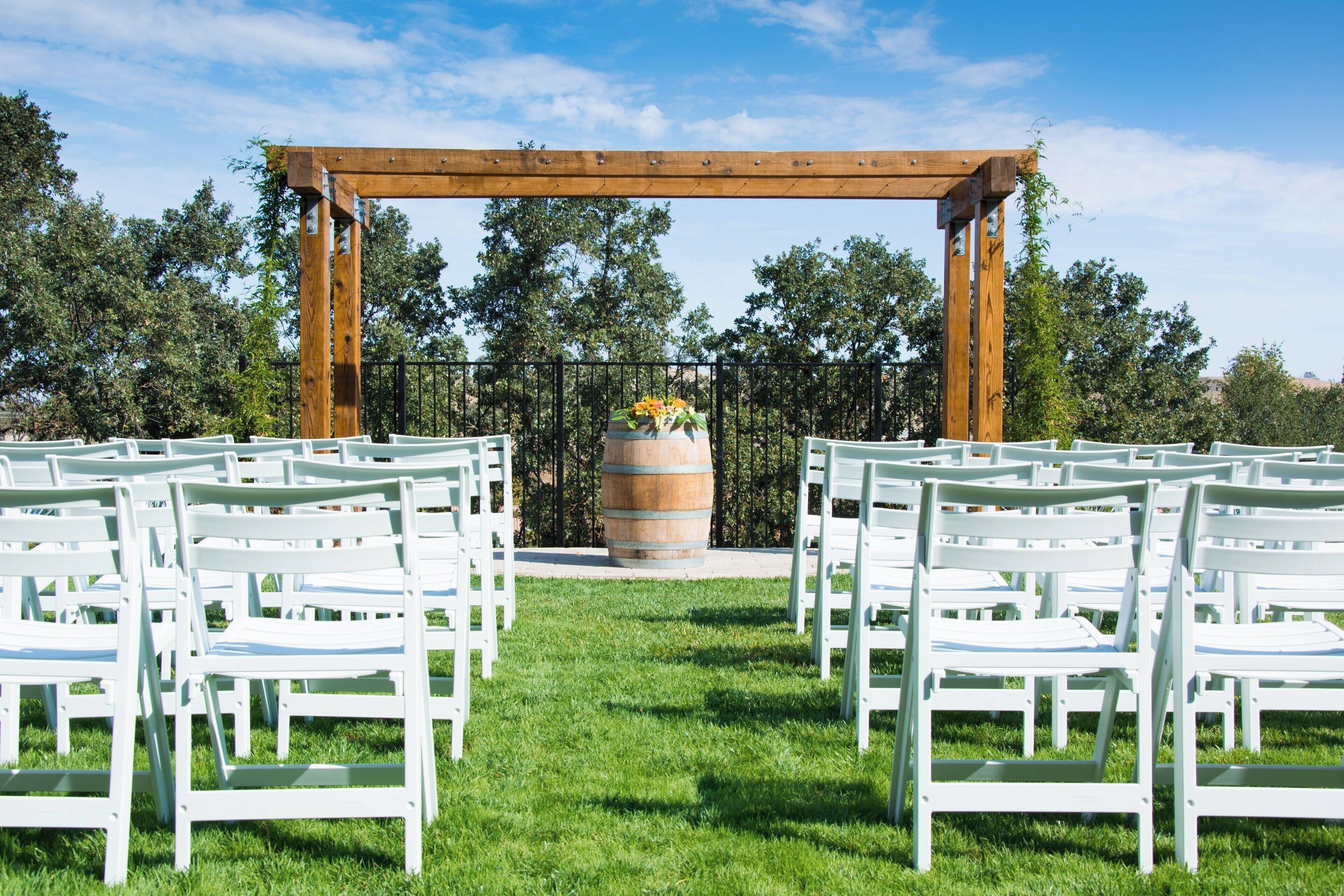 A wedding ceremony setup with white chairs arranged in rows under a wooden archway, with a barrel and flowers in the center.