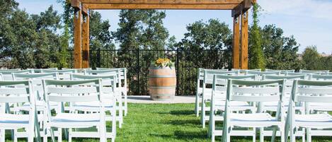 A wedding ceremony setup with white chairs arranged in rows under a wooden archway, with a barrel and flowers in the center.