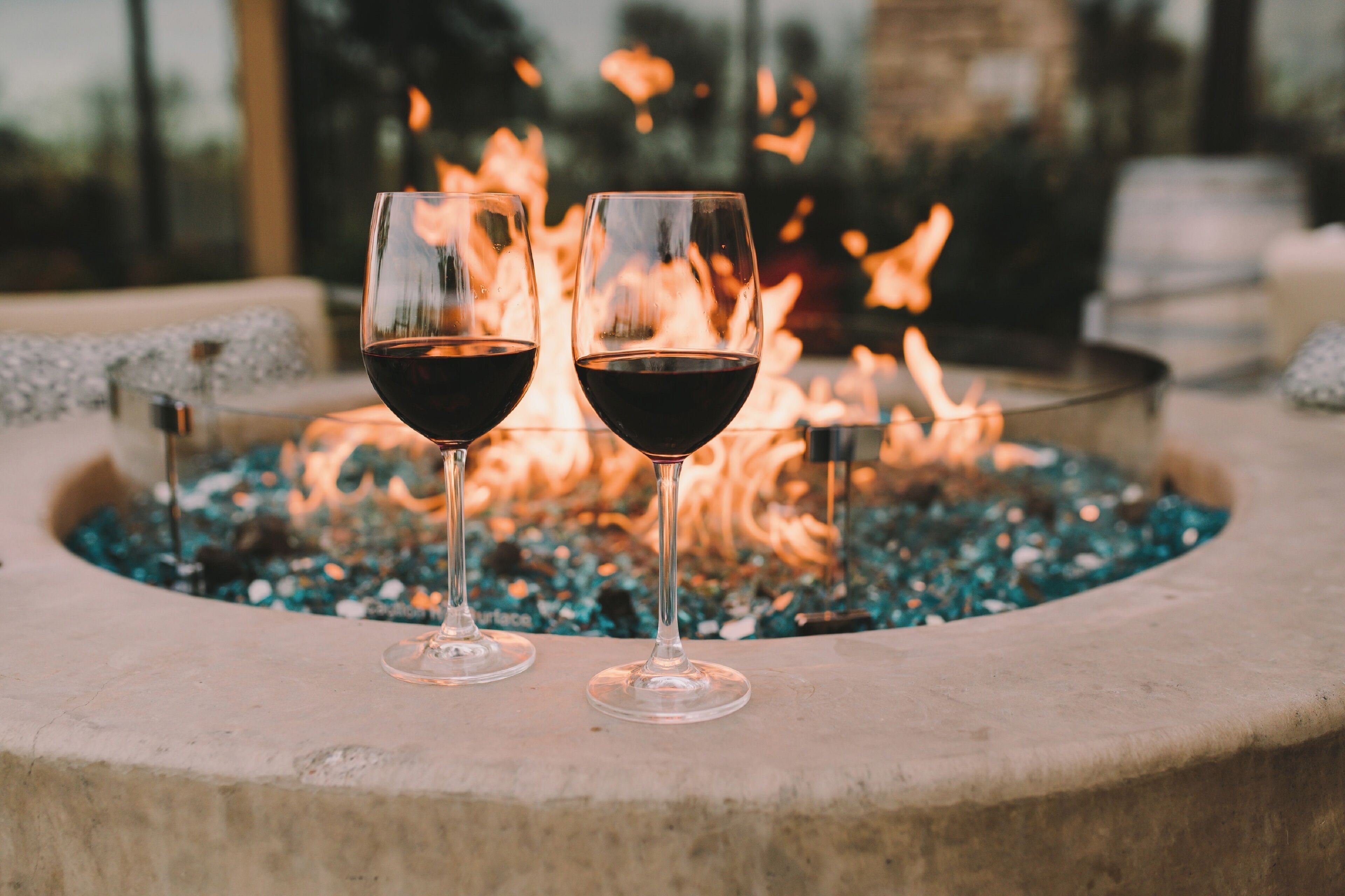 Deux verres à vin contenant un liquide rouge, posés sur une surface en pierre près d’un foyer.