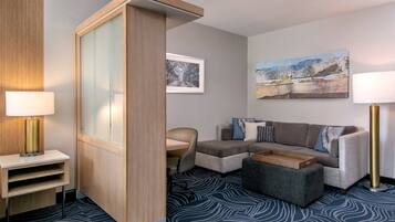 A hotel room with a sofa, a coffee table, a desk with a lamp, and a painting on the wall.
