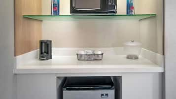 A compact kitchenette with a microwave, two water bottles, a coffee maker, a small refrigerator, and a glass shelf.