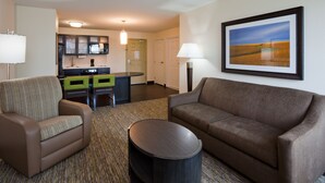 A hotel room with a brown sofa, a striped armchair, a small round table, a kitchenette with a green chair, and a framed landscape painting on the wall.