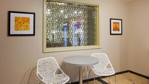 A room with a round table and two white chairs with intricate designs, a window with a decorative pattern, and two framed pictures on the walls.