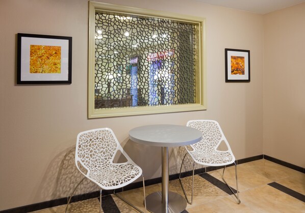 Une pièce comprenant une table ronde et deux chaises blanches ornées de motifs complexes, une fenêtre décorée d’un motif ornemental et deux tableaux encadrés accrochés aux murs.