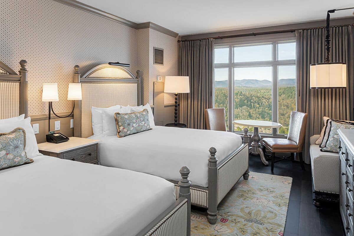A hotel room with two beds, a desk, a chair, a dresser, and a large window with a view.