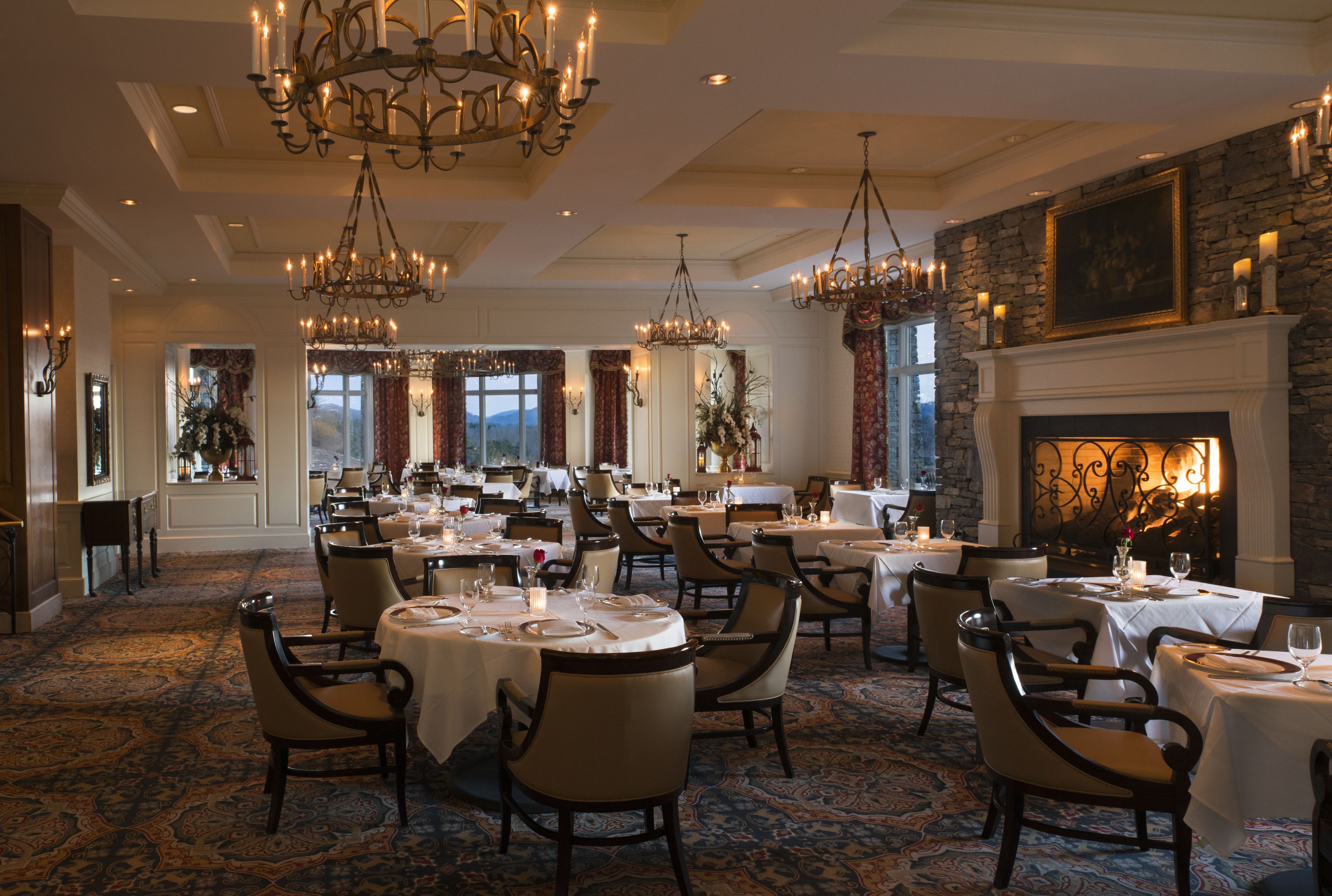 A spacious dining area with multiple round tables and chairs, chandeliers, and a fireplace.