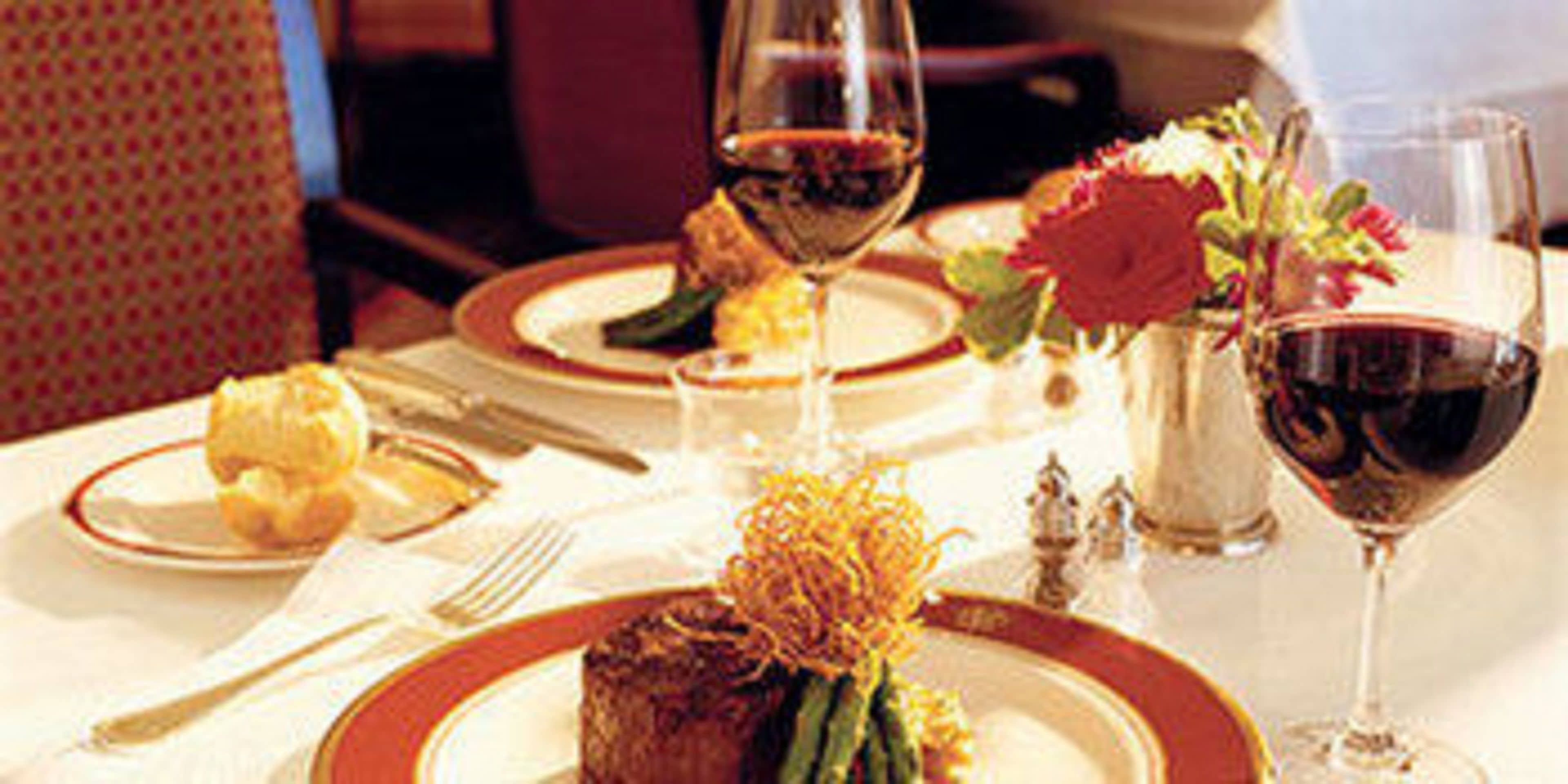 Una mesa de comedor dispuesta con platos, cubiertos, una copa de vino tinto y un centro de mesa con flores.