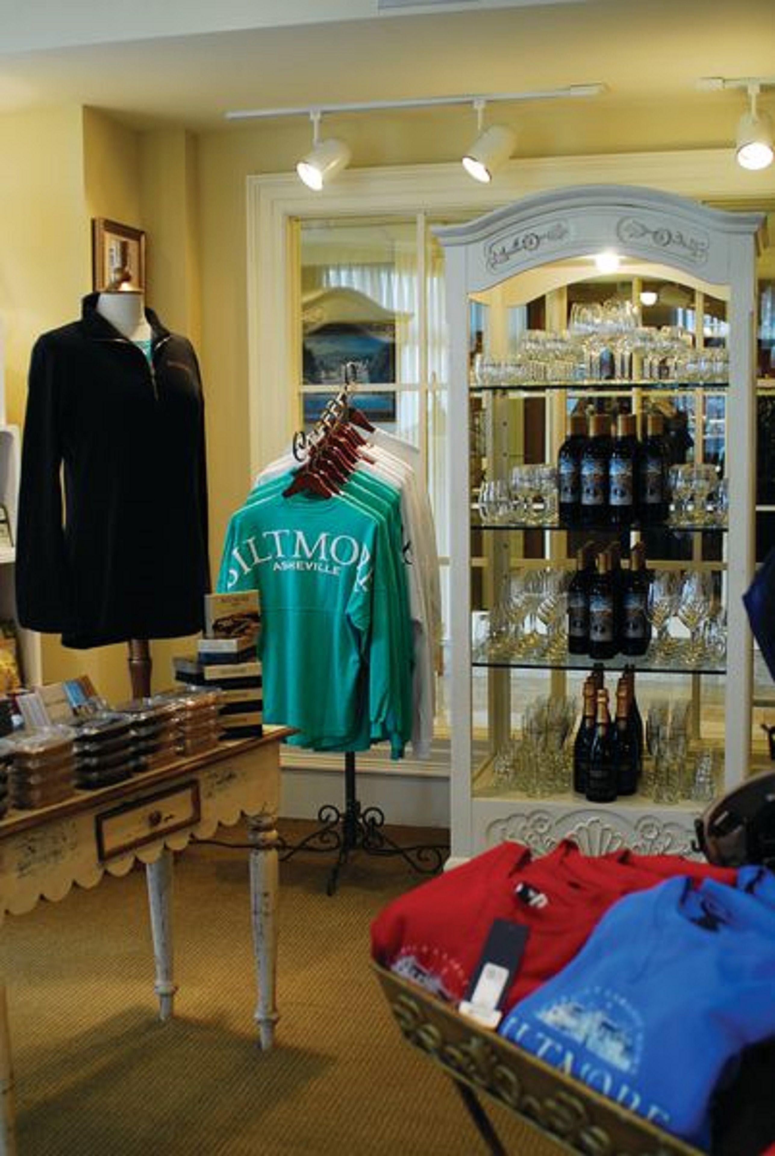Quarto de hotel boutique com um manequim exibindo uma camisa azul-turquesa, uma vitrine de vidro com garrafas e copos, e uma mesa antiga com um livro e uma pequena caixa.