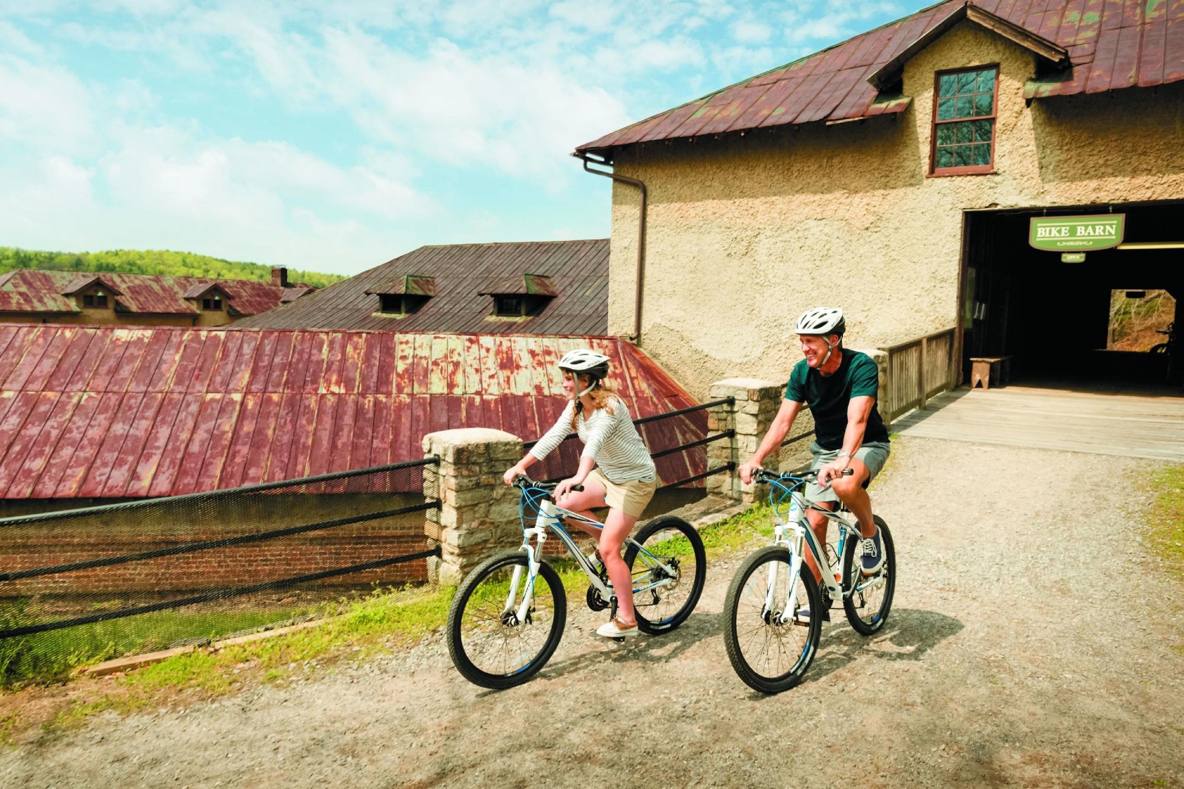 Deux personnes à vélo sur un chemin, avec un bâtiment rustique en arrière-plan.