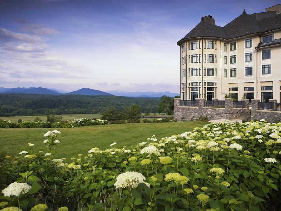 A large building with a stone facade and multiple windows, surrounded by lush greenery and a field of white flowers.