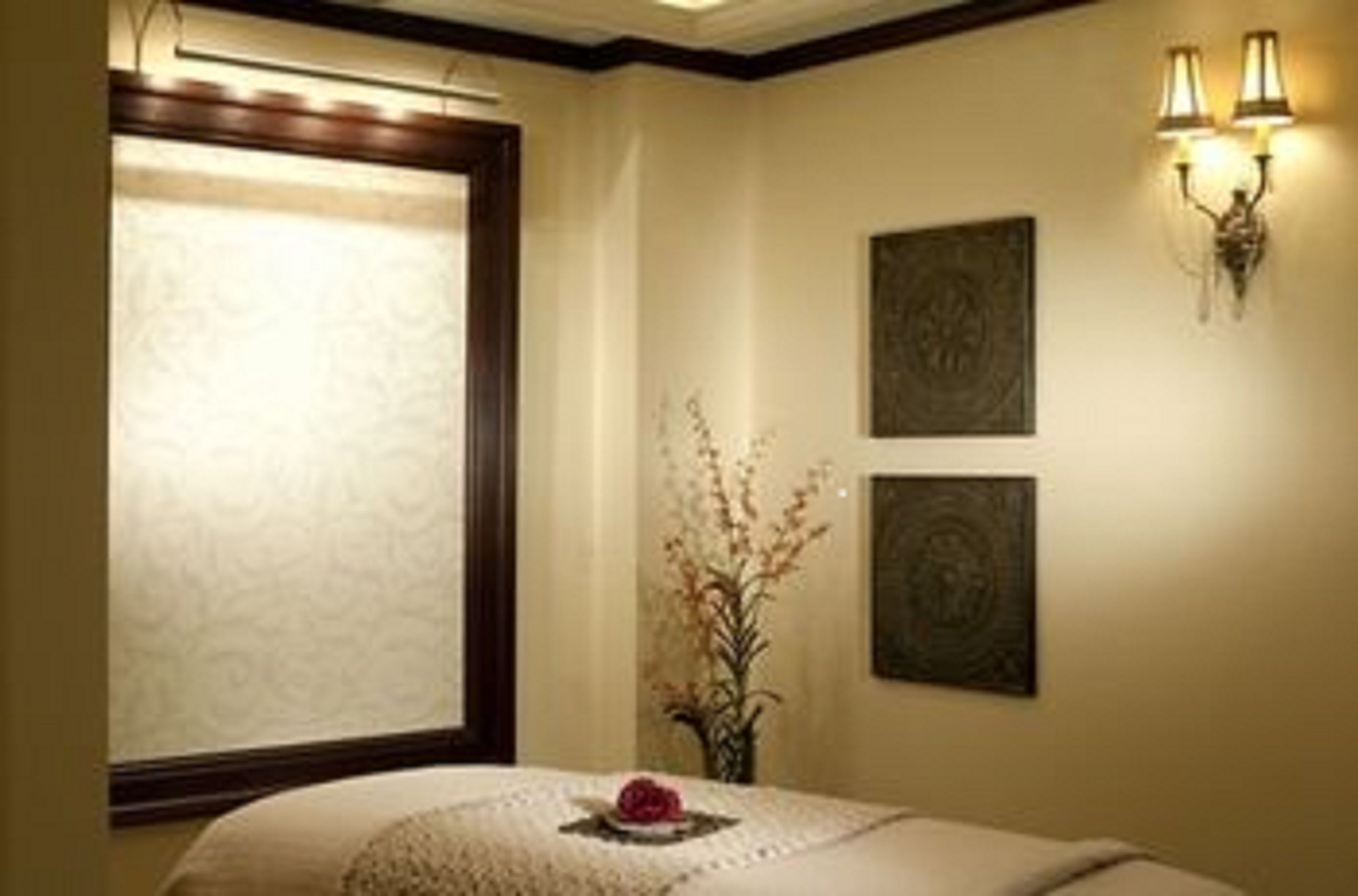A hotel room with a large window, a bed with a decorative pillow, a vase with flowers, and two wall-mounted lamps.