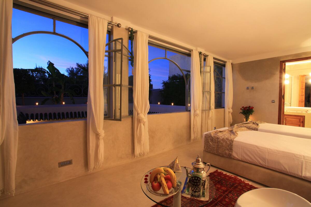 Una habitación de hotel con cama, una mesa con fruta, una lámpara decorativa y ventanas amplias con cortinas.