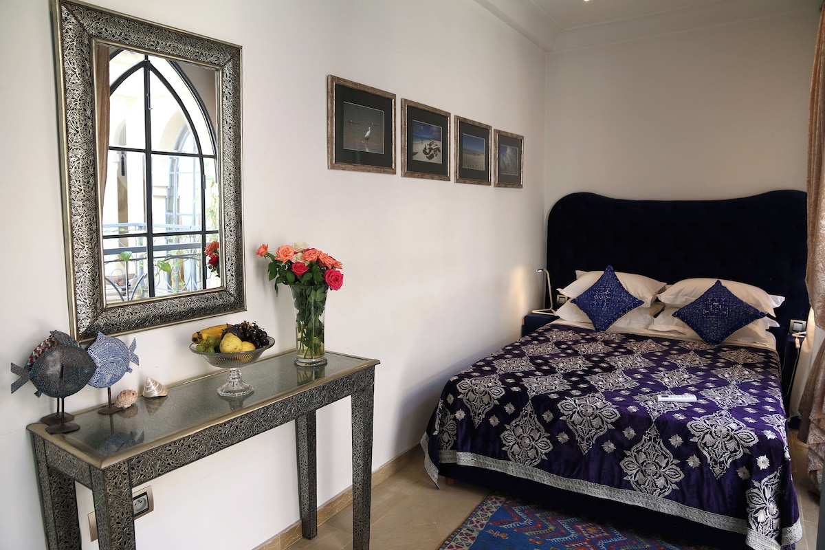 A bedroom with a large mirror, a bed with a patterned bedspread, a small table with decorative items, and framed pictures on the wall.