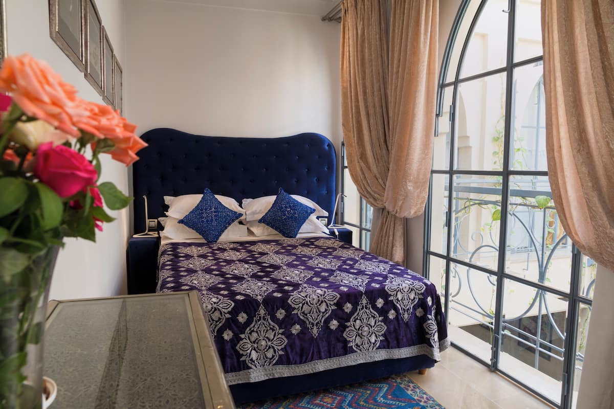 Un dormitorio con una cama grande, una mesita de noche, un florero con flores y vistas al exterior a través de una ventana.