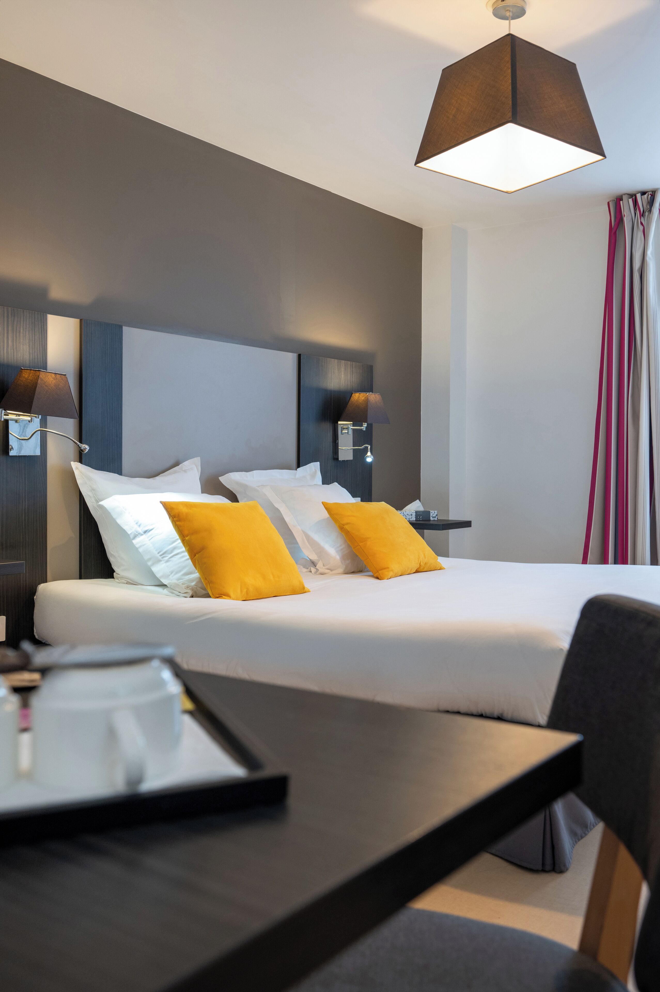 A hotel room with a bed, two bedside lamps, a table with a tray, and a hanging lamp.