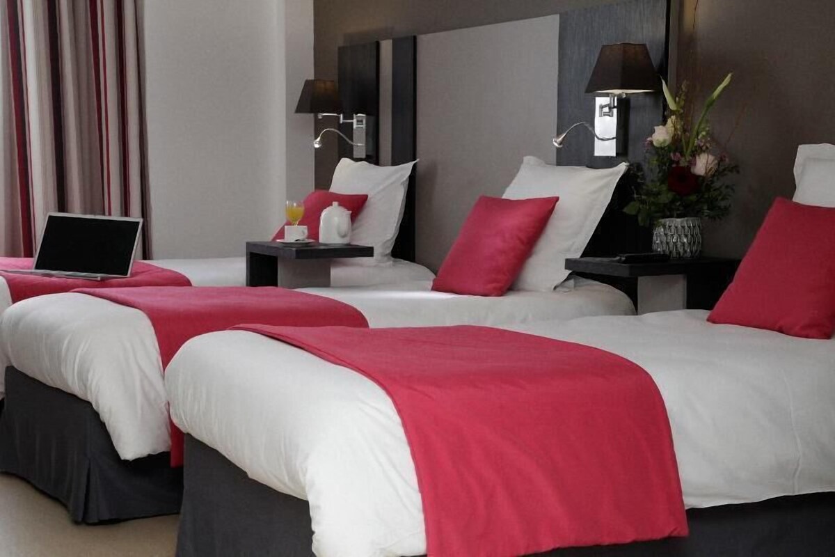 A hotel room with two beds, each with a red and white bedspread, bedside tables with lamps, and a laptop on one bed.