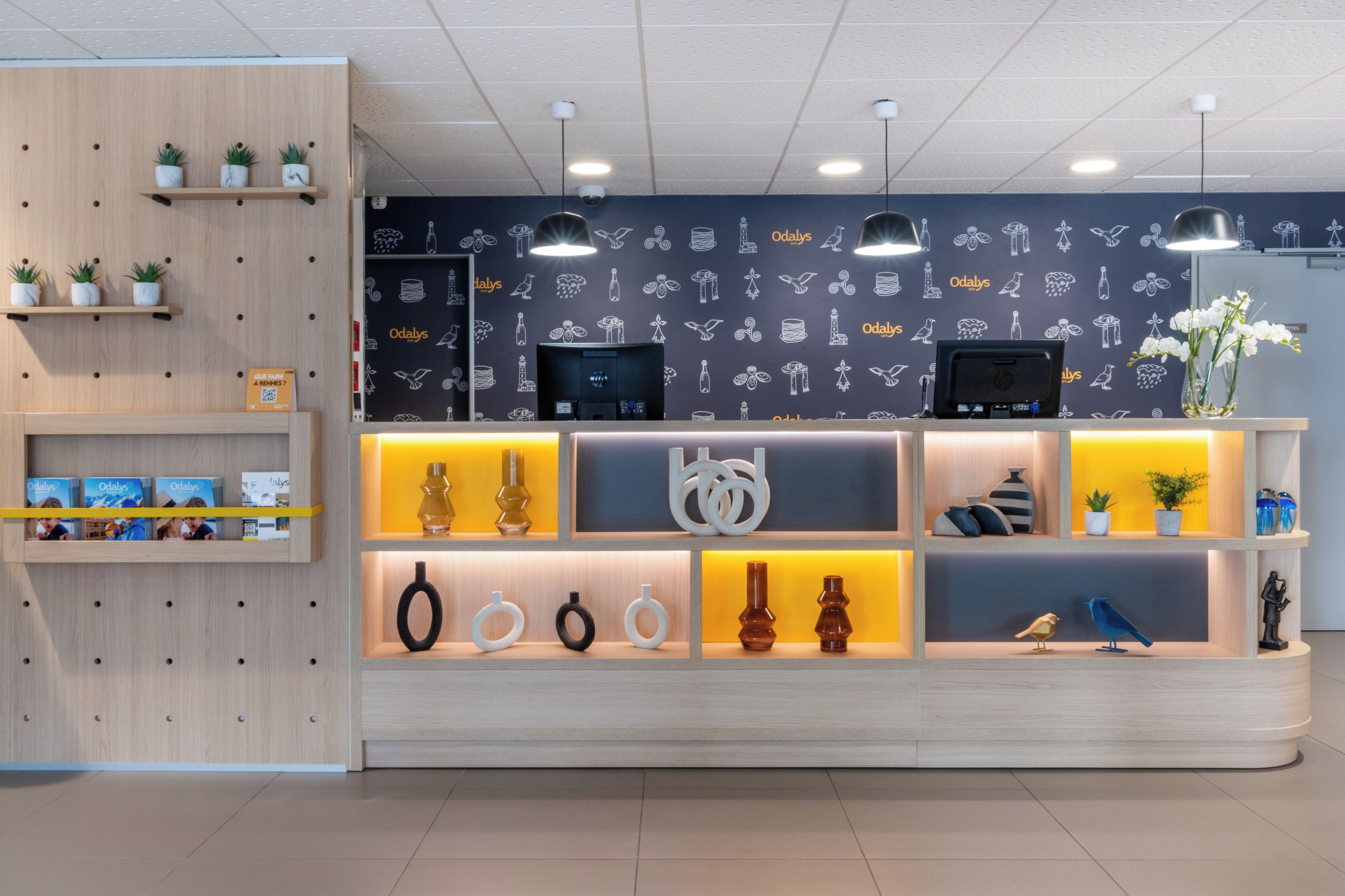 Un mostrador de recepción con diseño moderno, que incluye un panel de madera en la pared, un fondo oscuro con motivos y varios objetos decorativos en estantes.