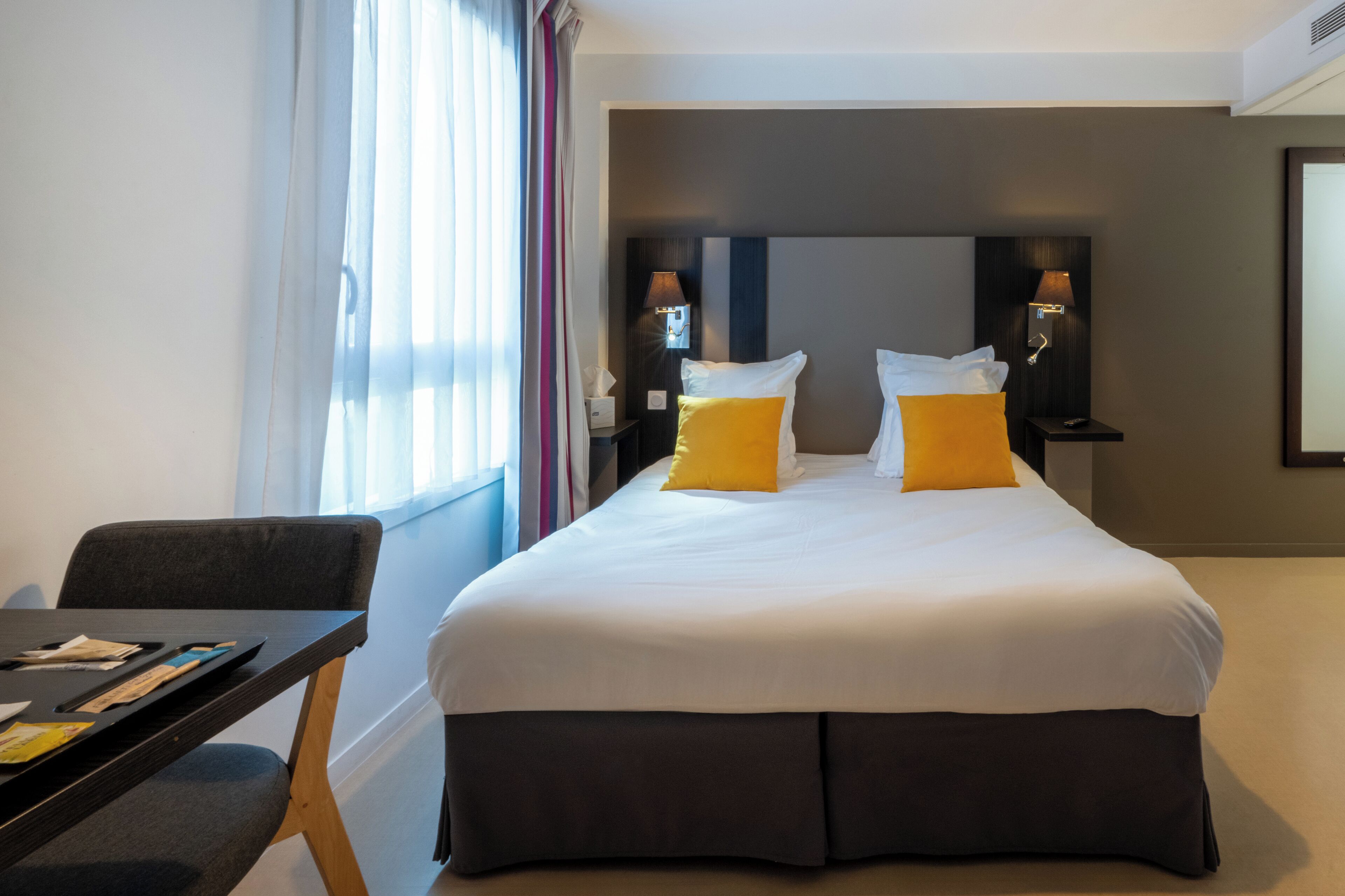 A hotel room with a large bed, two bedside lamps, a desk with a chair, and a window with curtains.