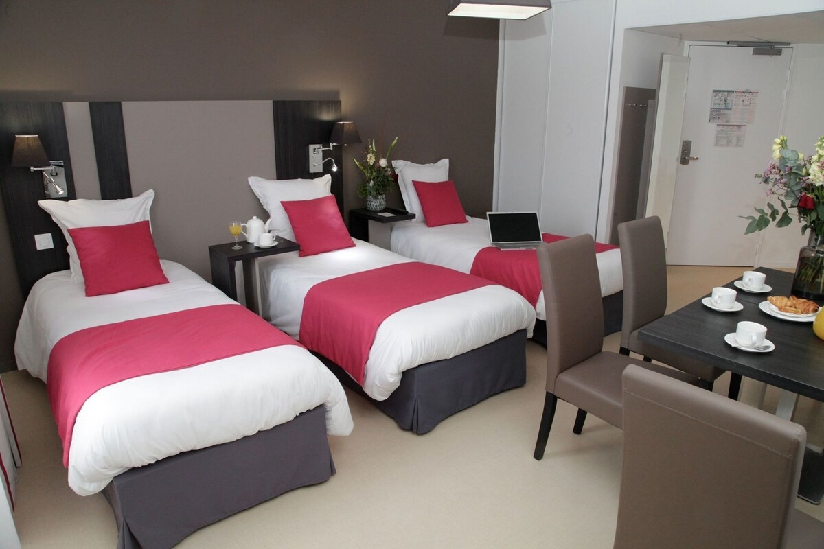 A hotel room with three beds, a dining table with chairs, a laptop on one bed, and a vase with flowers.