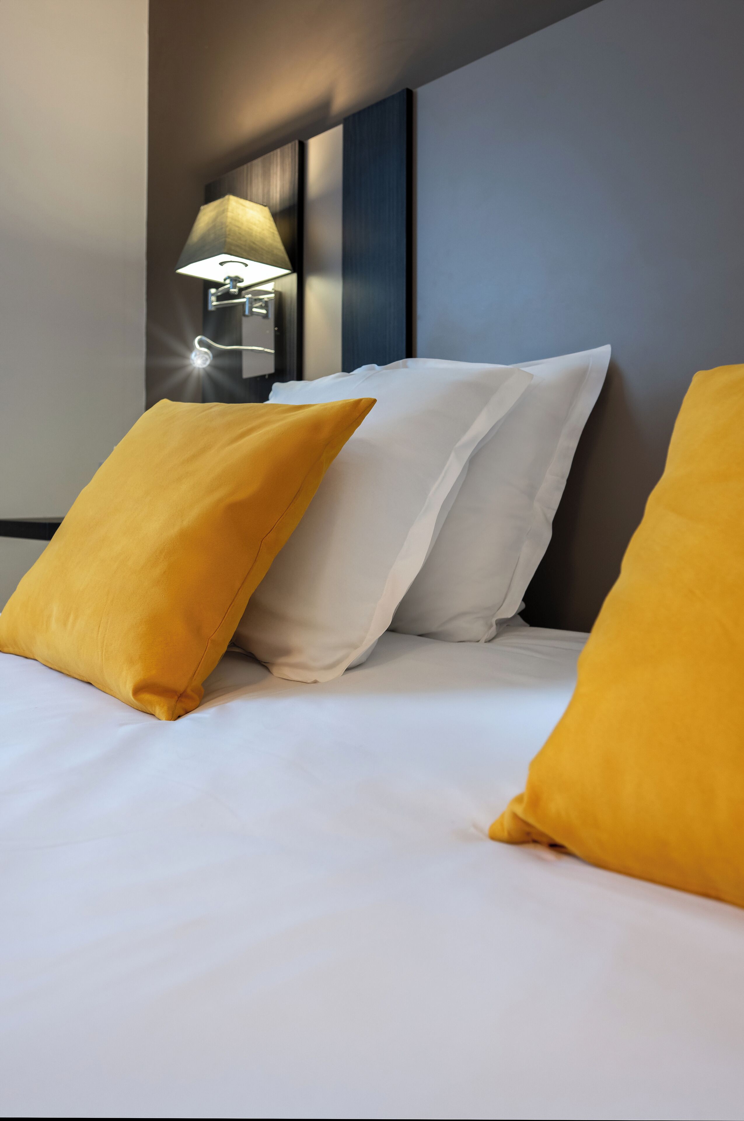 Una cama bien hecha con almohadas blancas y amarillas, una lámpara fijada a la pared y un cabecero oscuro.