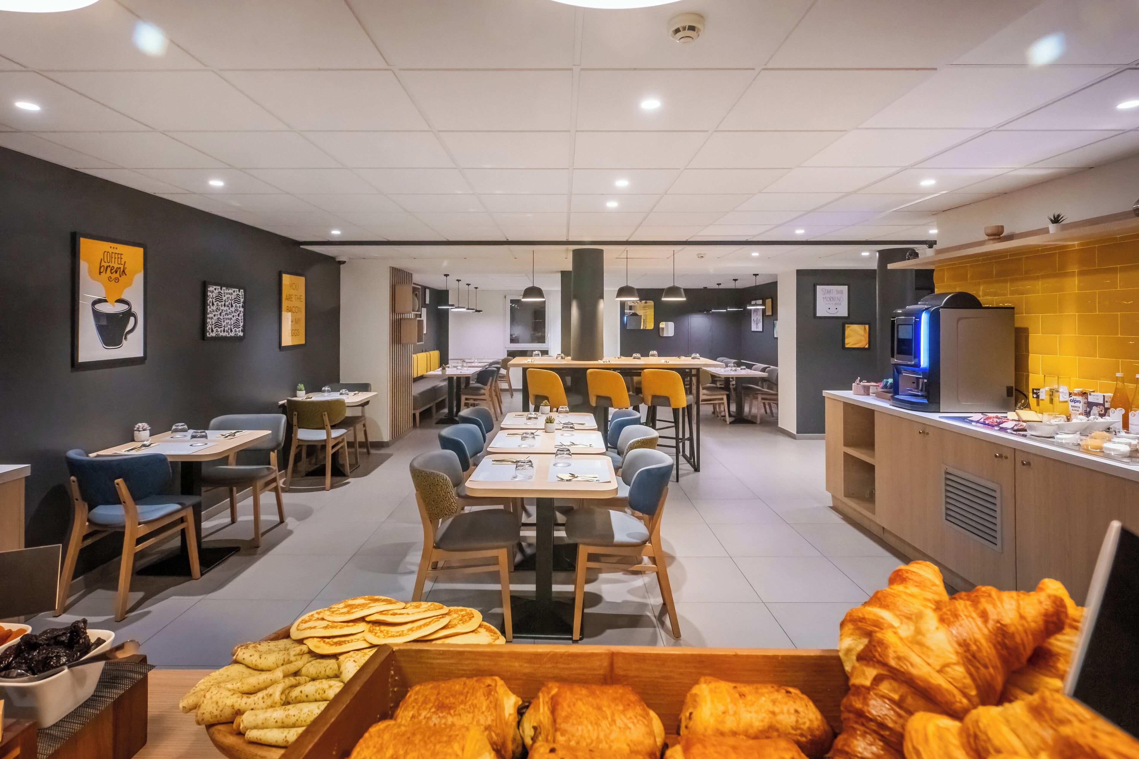 A modern cafe with multiple tables and chairs, a counter with a coffee machine, and a variety of pastries displayed in the foreground.