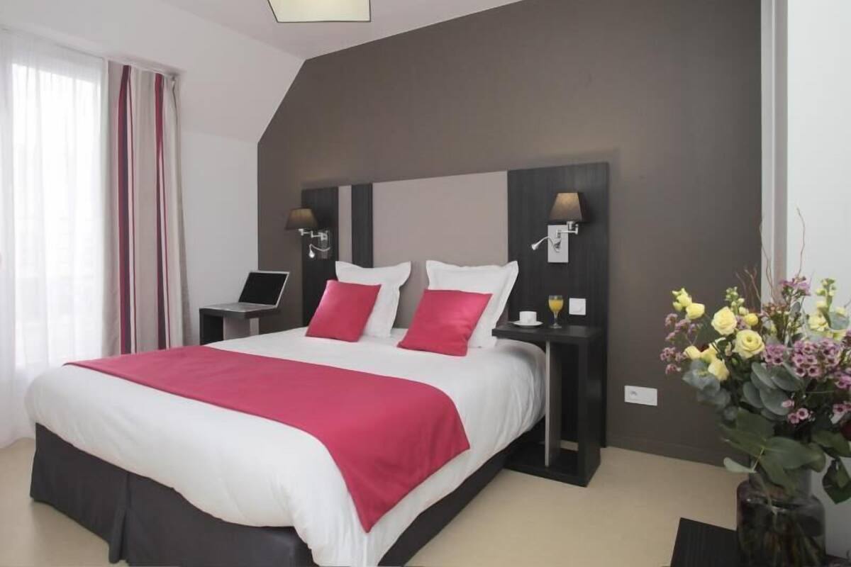 Une chambre d’hôtel moderne avec un grand lit, deux tables de chevet, un bureau, un ordinateur portable, un vase avec des fleurs et un luminaire au plafond.