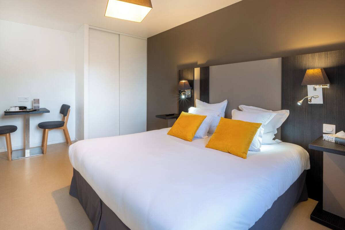 Une chambre d’hôtel moderne avec un grand lit, deux tables de chevet avec des lampes, un bureau avec deux chaises et une armoire.