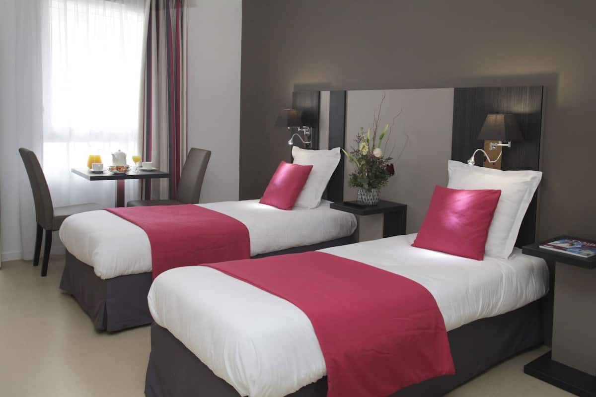 A hotel room with two beds, a dining table, chairs, a vase with flowers, and a magazine on a nightstand.