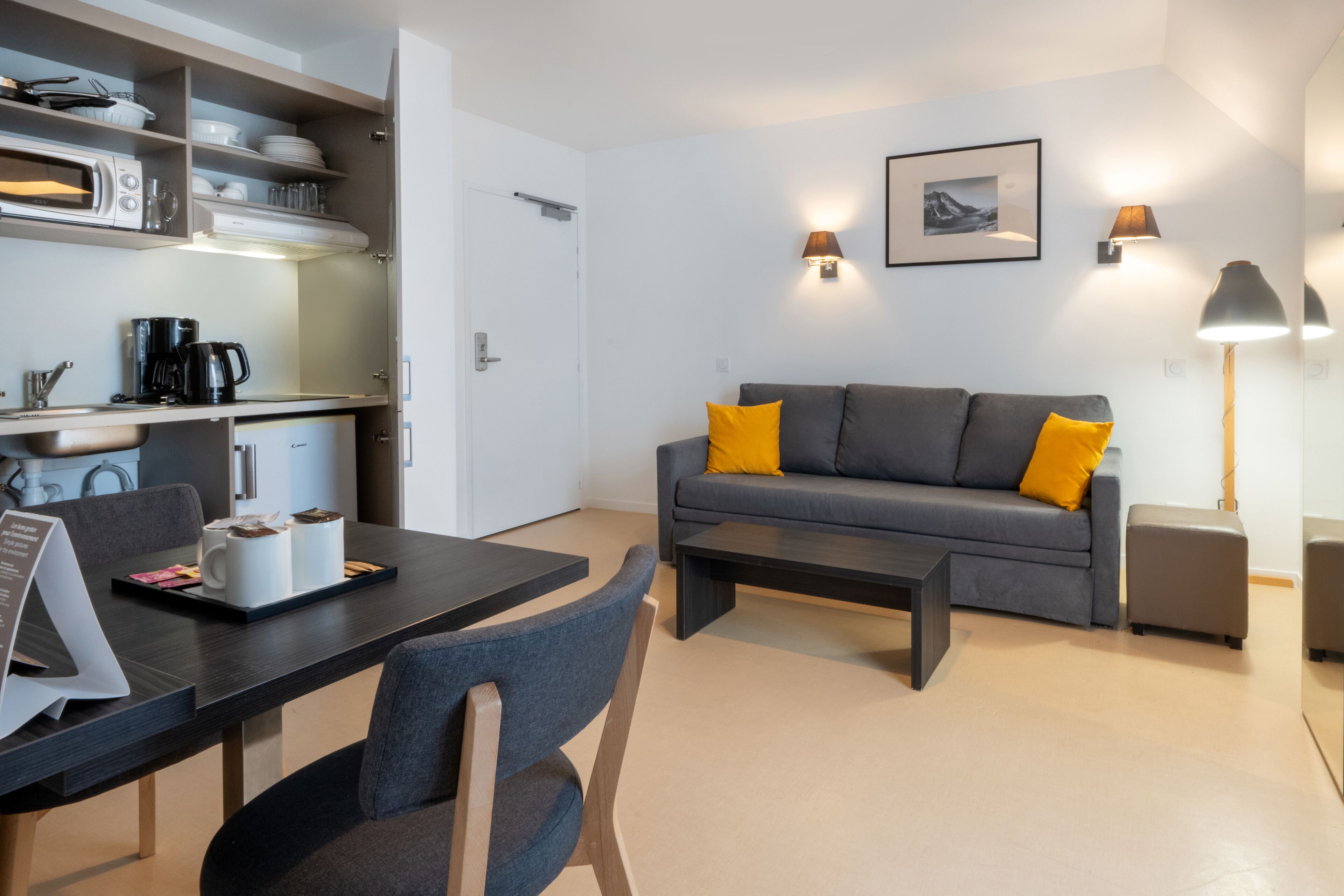 A modern living room with a grey sofa, yellow cushions, a coffee table, and a dining area with a laptop and coffee maker.